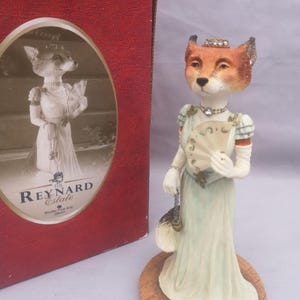 May include: A fox figurine in a period dress, holding a fan and a small purse. The fox wears a jewelled crown and necklace. The figurine stands on a wooden base, with a red box showing a similar image.