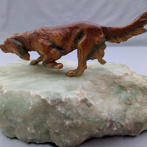 May include: A brown dog figurine in a crouching, alert stance, mounted on a large, irregular, light green stone base. The dog has textured fur details and is positioned as if tracking or hunting, with its head lowered and tail extended.