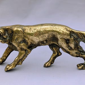 Könnte beinhalten: Eine goldfarbene Metallskulptur eines Hundes in Gehpose. Der Hund hat einen langen Körper, einen langen Schwanz und Schlappohren. Die Skulptur ist detailliert, mit sichtbarem Fell und Muskeldefinition. Der Hund ist etwa 15 cm lang.