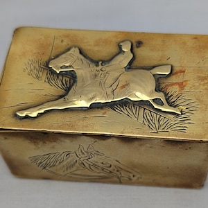 Puede incluir: Una caja rectangular de latón con un relieve de un jinete sobre un caballo al galope en la tapa. El frente de la caja muestra una imagen grabada de una cabeza de caballo. La superficie metálica presenta un tono dorado pulido con pátina.