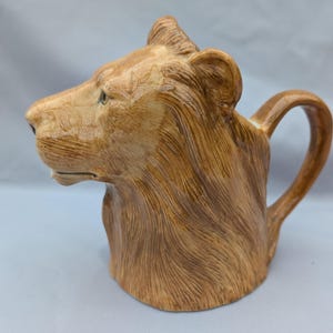 Könnte beinhalten: Ein Keramikkrug in Form eines Löwenkopfes mit detaillierter Mähne und Gesichtszügen. Der Krug ist hauptsächlich braun mit helleren Akzenten und hat einen gebogenen Griff.