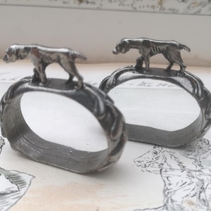 May include: Two silver-toned napkin rings. Each ring features a detailed sculpture of a dog on top. The rings have a textured, braided design. The background includes a vintage paper with a drawing.