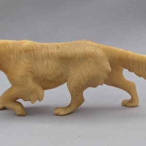 Puede incluir: Figura detallada de perro de color claro en posición de caminar. La escultura presenta pelaje texturizado y una cola larga y fluida. Las orejas del perro son caídas y su cuerpo está ligeramente encorvado, dándole una apariencia realista. La figura es probablemente de resina.