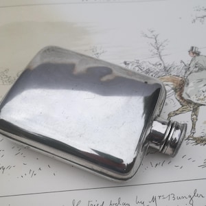 Puede incluir: Una petaca de metal plateado pulido con forma rectangular y esquinas redondeadas. La petaca cuenta con un tapón de rosca en el lateral y una superficie lisa y reflectante que captura la luz y los reflejos del entorno en todo su cuerpo.