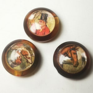 Puede incluir: Tres botones redondos de resina marrón con imágenes de personas y animales. El primer botón tiene una imagen de una persona con un abrigo rojo y un sombrero de copa. El segundo botón tiene una imagen de dos personas en un entorno boscoso. El tercer botón tiene una imagen de la cabeza de un caballo.