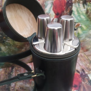 May include: A green leather carrying case with a zip closure and a shoulder strap. The case contains three silver shot glasses.