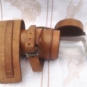 May include: A brown leather flask with a glass bottle and a silver cap. The flask has a strap and buckle closure.