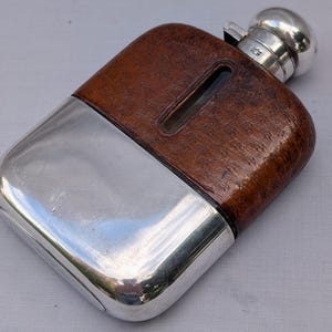 Può includere: Una fiaschetta in argento con un rivestimento in pelle marrone sulla parte superiore e una finestrella verticale. La fiaschetta presenta un tappo argentato arrotondato e una base in argento lucido, mostrando una combinazione di metallo e pelle.