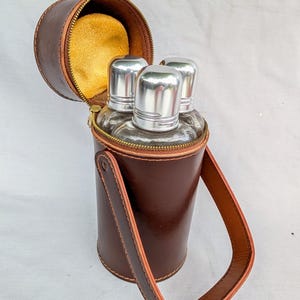 Può includere: Un set di fiaschette in pelle marrone con tre bottiglie di vetro e tappi argentati. La custodia cilindrica ha una parte superiore con cerniera e un manico. L'interno è foderato in tessuto giallo. Il set di fiaschette è progettato per trasportare liquidi.