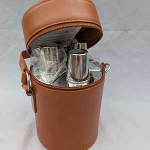 May include: A brown leatherette travel bar set in a cylindrical case with a zipped top. The case contains several stainless steel cups. The set includes a handle and a shoulder strap attachment.