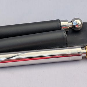 May include: Two stainless steel flasks with spherical tops, one silver and one gold, are in black leather sleeves. The flasks are held in a black leather carrying case with a flap. The background is a light, neutral colour.