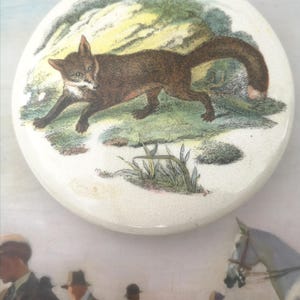 Pode incluir: Um azulejo de cerâmica redondo com uma imagem pintada à mão de uma raposa correndo por um campo de grama. A raposa é marrom com o peito branco e uma cauda espessa. O fundo é verde claro e amarelo.