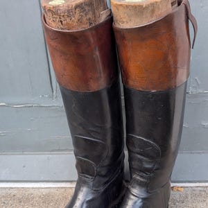以下が含まれることがあります： 上部に茶色のレザーカフが付いた、背の高い黒いレザーの乗馬ブーツ一足。各ブーツには、レザーの形状を維持するために、丸いハンドルが付いた木製のシューツリーがシャフトに挿入されています。