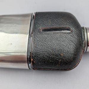 Puede incluir: Un frasco de cadera vintage con una parte superior de metal plateado y un cuerpo cubierto de cuero negro. El frasco tiene una forma redondeada con un pequeño pico cilíndrico. El cuero tiene una apariencia texturizada.