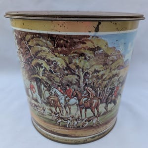 May include: Vintage metal container with a lid. The container features a detailed illustration of a fox hunt scene with riders on horseback, hounds, and a forest backdrop. The colors include shades of brown, green, and blue.