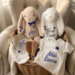May include: A wicker basket holds a collection of baby items. Two plush bunny rabbits, one beige and one white, are nestled among a white onesie, comb, mittens, and a drawstring bag. All items are embroidered with blue text and star accents.