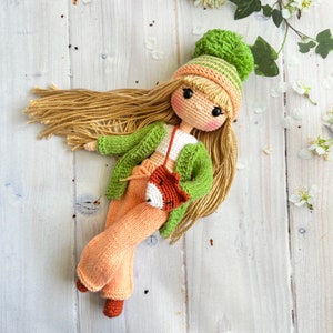 May include: A crocheted doll with long blonde hair wearing a green cardigan, peach trousers, and a green and peach hat. The doll is holding a small crocheted fox.