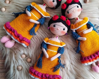 May include: Three crocheted dolls with black hair, wearing yellow dresses with colorful accents and blue scarves. The dolls have red flowers in their hair and are arranged on a white fluffy surface.