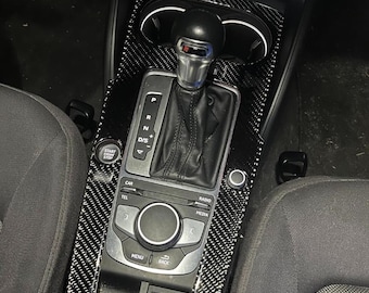 Puede incluir: Kit de molduras interiores de fibra de carbono para la consola central de un coche. El kit incluye una palanca de cambios, controles multimedia y portavasos. El embellecedor tiene un patrón de cuadros blancos y negros. El botón de arranque/parada del coche es visible.