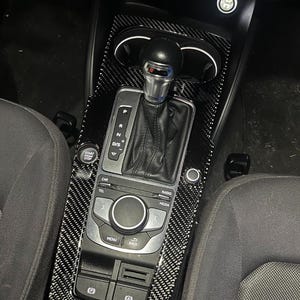 Puede incluir: Kit de molduras interiores de fibra de carbono para la consola central de un coche. El kit incluye una palanca de cambios, controles multimedia y portavasos. El embellecedor tiene un patrón de cuadros blancos y negros. El botón de arranque/parada del coche es visible.