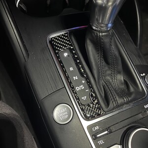 Può includere: Primo piano dell'interno di un'auto, che mostra il pannello del cambio con un design in fibra di carbonio. Il pannello mostra le posizioni del cambio: P, R, N, D/S e un pulsante start/stop. La leva del cambio è in pelle nera.