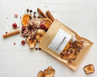 May include: A brown paper bag with a white label that says "Glimmering Potpourri" is spilling out dried fruit, cinnamon sticks, star anise, coffee beans, and other spices onto a white surface.