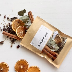 May include: A kraft paper bag of potpourri with dried orange slices, cinnamon sticks, and star anise. The bag has a label that reads "Simmering Potpourri". Scattered around are additional ingredients like cloves and eucalyptus.