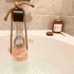 May include: A pink Himalayan salt block is suspended in a bathtub by a rope and wooden dowel. The salt block is being dissolved by running water from a faucet. A jar of bath salts labeled "Vanilla Rose Hibiscus Salt Soak" is on the edge of the tub.
