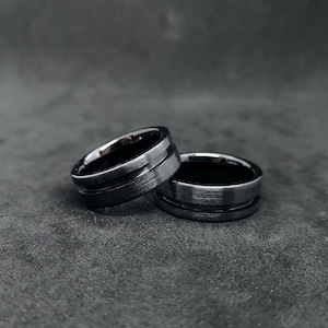 Könnte beinhalten: Zwei schwarze Ringe aus Wolframcarbid mit gebürsteter Oberfläche und einem gerillten Design.