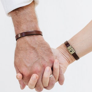 May include: A couple holding hands, each wearing a brown leather bracelet. The woman's bracelet has a small metal charm with the word "love" engraved on it.