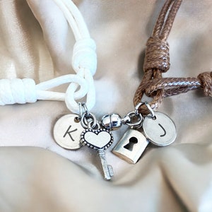 Könnte beinhalten: Zwei passende Freundschaftsarmbänder mit einem Schlüssel- und Schlossanhänger, einem Herzanhänger und personalisierten Buchstabenanhängern. Die Armbänder sind aus weißem und braunem Kordel.