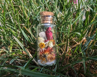 May include: A clear glass bottle with a cork stopper filled with dried flowers and a white crystal. The bottle is sitting on a bed of green grass.