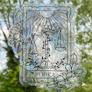 May include: A holographic sticker featuring a black and white illustration of the Libra zodiac sign, a scale with a sun above it, and the word "LIBRA" below. The sticker is surrounded by black and white floral designs.