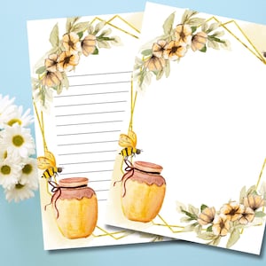 Può includere: Due fogli di carta da lettere con un design ad acquerello. Il design presenta vasetti di miele, api e composizioni floreali con una cornice geometrica dorata. Un foglio ha carta a righe, l'altro è vuoto. Un vaso di margherite è a sinistra.