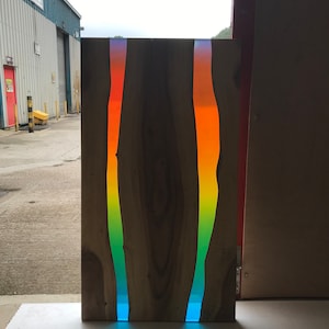 May include: A wooden table top with two vertical rainbow resin inlays. The resin is a gradient of blue, green, yellow, orange, and red. The table top is standing on a white surface.