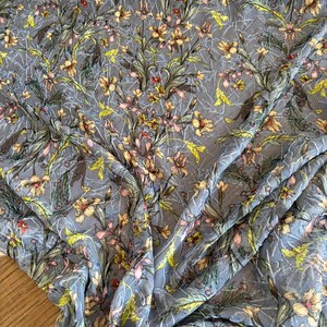 Può includere: Un tessuto leggero e trasparente con uno sfondo blu ardesia e un motivo floreale denso e intricato in tonalità di giallo, verde tenue e rosa delicato. Il materiale presenta una sottile consistenza stropicciata su tutta la superficie.