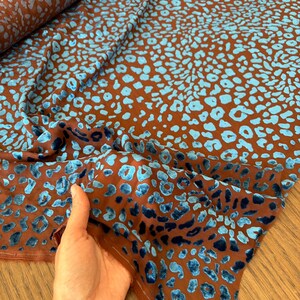 Könnte beinhalten: Ein Stoffballen mit brauner Grundfarbe und einem sich wiederholenden Leopardenmuster in leuchtendem Hellblau. Das Material hat eine strukturierte, samtartige Oberfläche, bei der die blauen Flecken vom glatten braunen Hintergrund abheben.