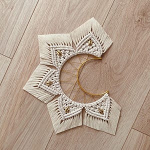 Può includere: Una decorazione da parete in macramè con un design a corda annodata color bianco sporco disposta a raggiera attorno a una mezzaluna in metallo dorato. L'oggetto presenta perline metalliche dorate inserite nelle sezioni in fibra di cotone intrecciata.
