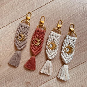 Op de afbeelding: Vier macramé sleutelhangers met geknoopt koord in beige, terracotta en twee tinten gebroken wit. Elke sleutelhanger bevat een goudkleurige metalen karabijnhaak, een ring en een centrale zon- en maansikkelbedel met steraccenten, afgewerkt met een kwastje.