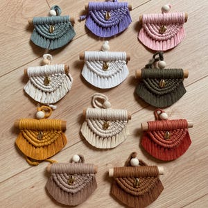 May include: A selection of handmade macrame purses in shades of teal, purple, pink, white, olive green, mustard yellow, beige, and rust. Each purse has a wooden dowel, a wooden bead, and a gold-coloured clasp. The purses are displayed on a light wood surface.