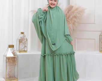 May include: A young person wearing a light green dress with a ruffled hem and a matching headscarf. The dress has long sleeves and a modest neckline. The person is smiling and looking at the camera.