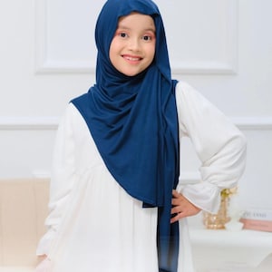 May include: A young girl in a navy blue hijab and a white dress. The hijab covers her head and shoulders. The dress has long sleeves and a flowing skirt. The girl is smiling. The image is well-lit and the colours are vibrant.