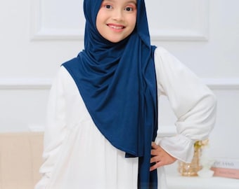 May include: A young girl wearing a navy blue hijab and a white dress. The hijab drapes over her head and shoulders, framing her face. The dress has long sleeves and a flowing skirt. The girl is smiling and looking at the camera.