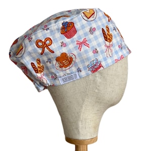 May include: A scrub cap featuring a light blue and white gingham pattern with a print of various baked goods including muffins, pretzels, pancakes, and baguettes, along with small pink bows. A white label reads SCRUBBED UP in black text.