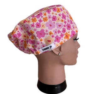 May include: A scrub cap with a pink, orange, and white floral pattern. The cap is displayed on a mannequin head. The text "SCRUBBED UP" is printed on a white tag on the cap.