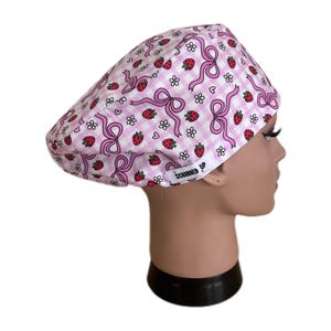 May include: A pink and white scrub cap with a gingham pattern, decorated with pink bows, strawberries, and white flowers. The cap is designed for medical use. The text "SCRUBBED UP" is visible on the side.