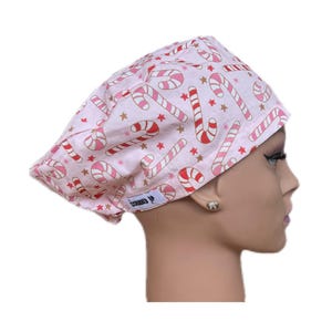 May include: A light pink scrub cap with a repeating pattern of red and white candy canes and gold stars. The cap has a curved design and is made of a soft fabric. The tag on the cap says "SCOOBED UP".