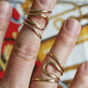 K&ouml;nnte beinhalten: Zwei goldfarbene Ringe, jeder mit einem einzigartigen, geschwungenen Design. Die Ringe werden an zwei Fingern einer Hand getragen.