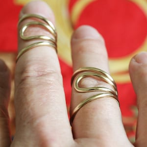 K&ouml;nnte beinhalten: Zwei goldene Ringe mit einem einzigartigen, abstrakten Design. Die Ringe werden am Zeige- und Mittelfinger einer Hand getragen.