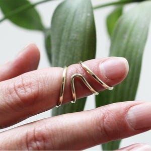 May include: A gold-colored, minimalist ring wraps around a finger. The ring features a simple, elegant design with a curved and linear shape. The jewelry is shown against a soft, blurred background of green leaves.